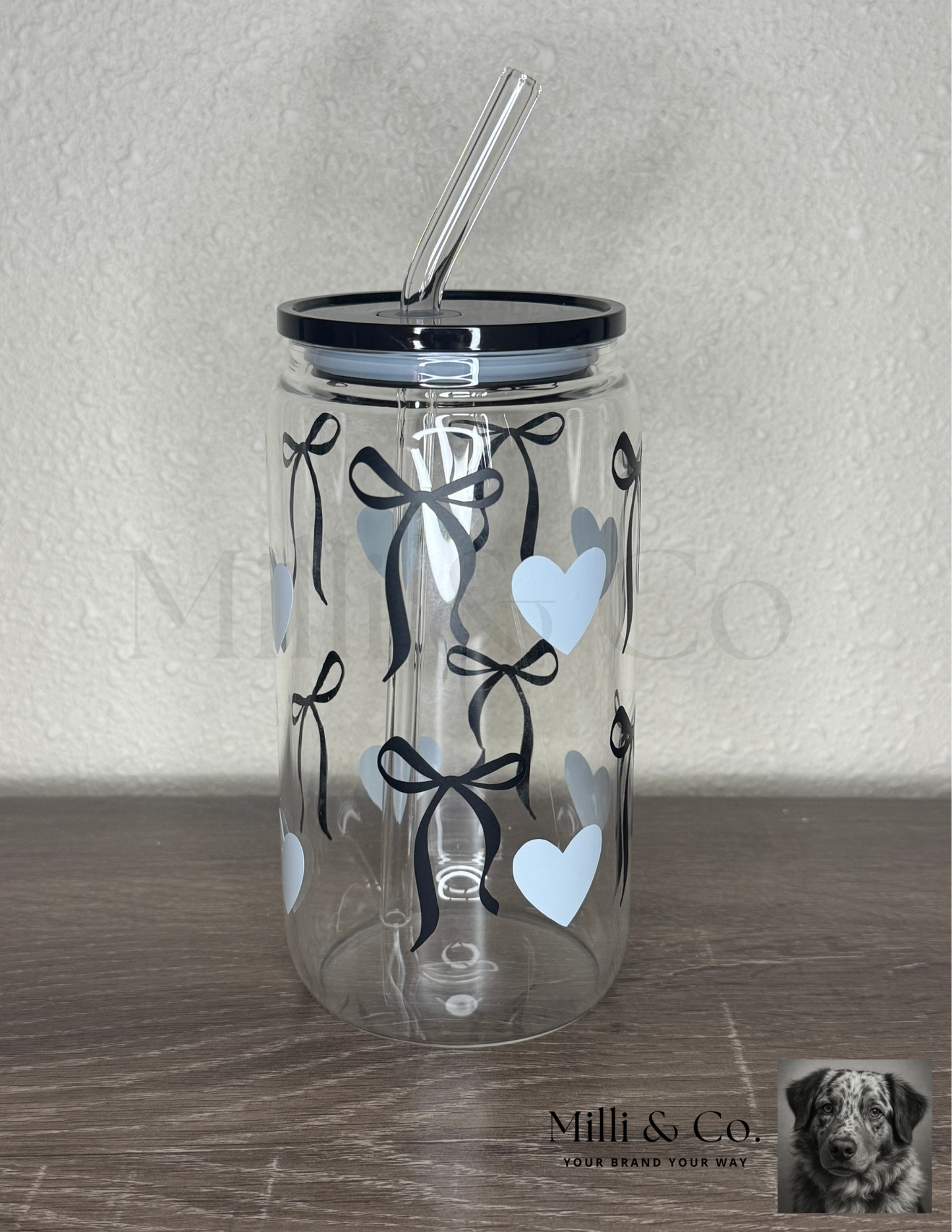 20oz clear glass tumbler with reusable glass straw, black bow and blue heart design, plastic lid, displayed on wooden surface.