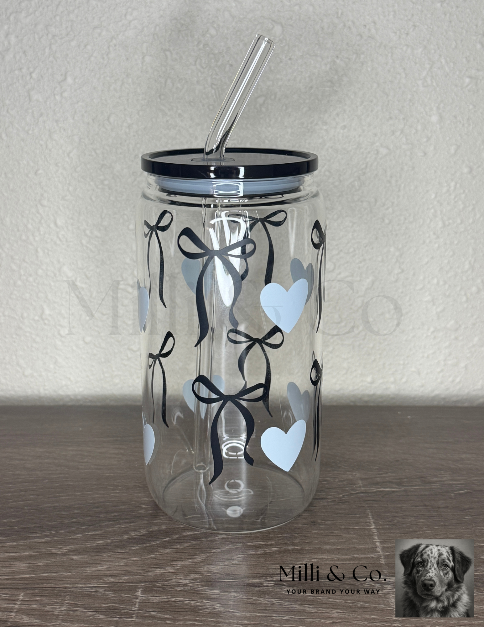20oz clear glass tumbler with reusable glass straw, black bow and blue heart design, plastic lid, displayed on wooden surface.